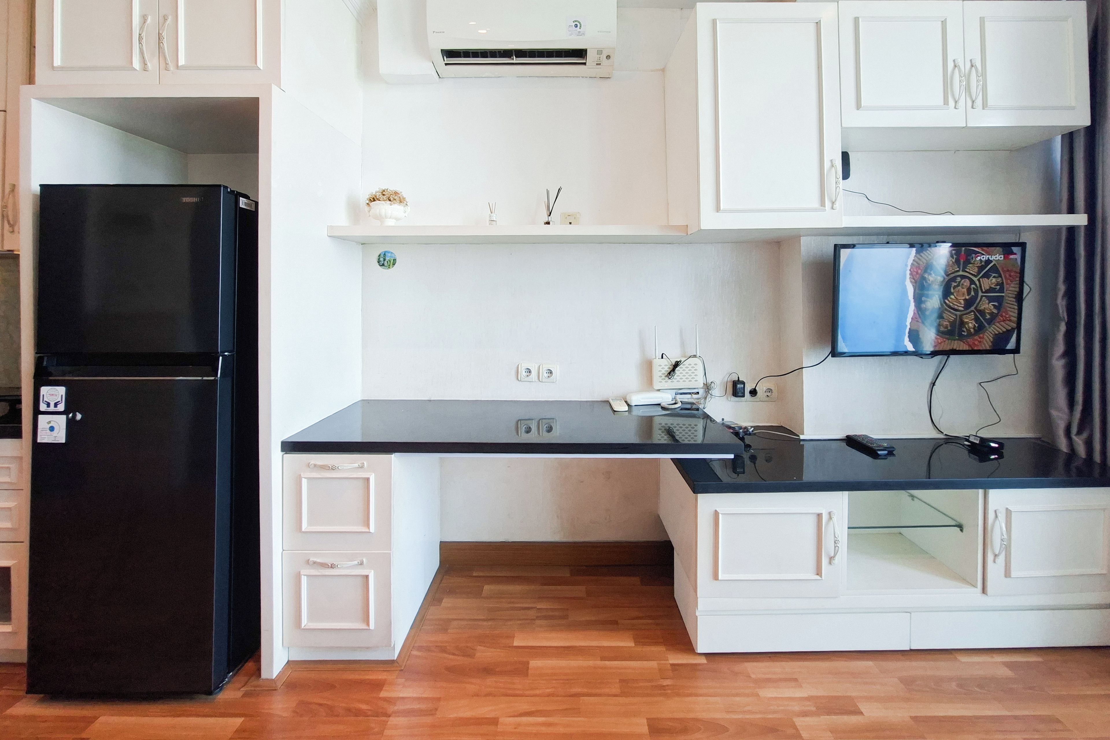 Cocina compacta con un refrigerador negro, armarios blancos, encimera negra y un televisor montado en la pared.