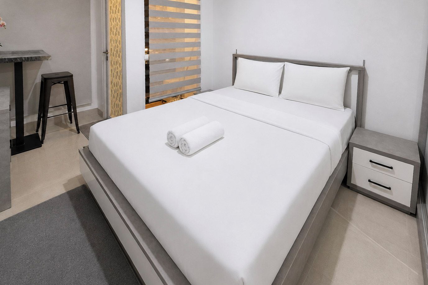 A modern hotel room with a large bed, a small bedside table with drawers, a desk with a chair, and a sliding door with a patterned curtain.