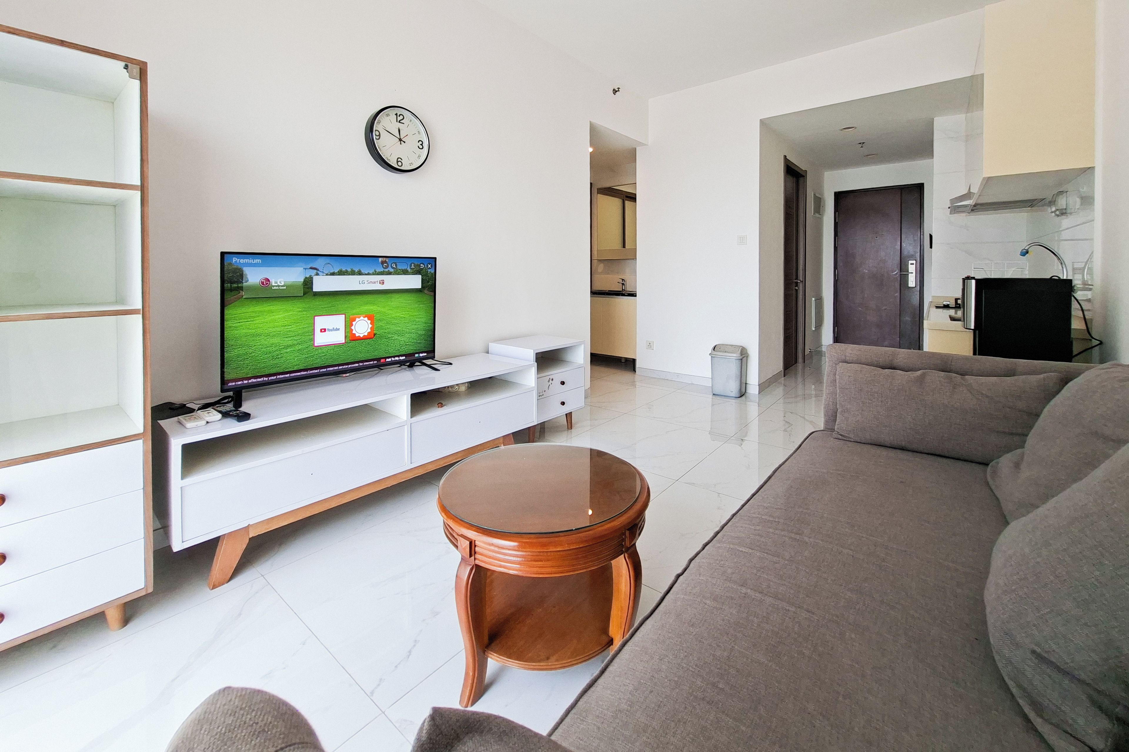 A living room with a flat-screen TV on a white stand, a round wooden coffee table, a grey sofa, and a kitchen area in the background.