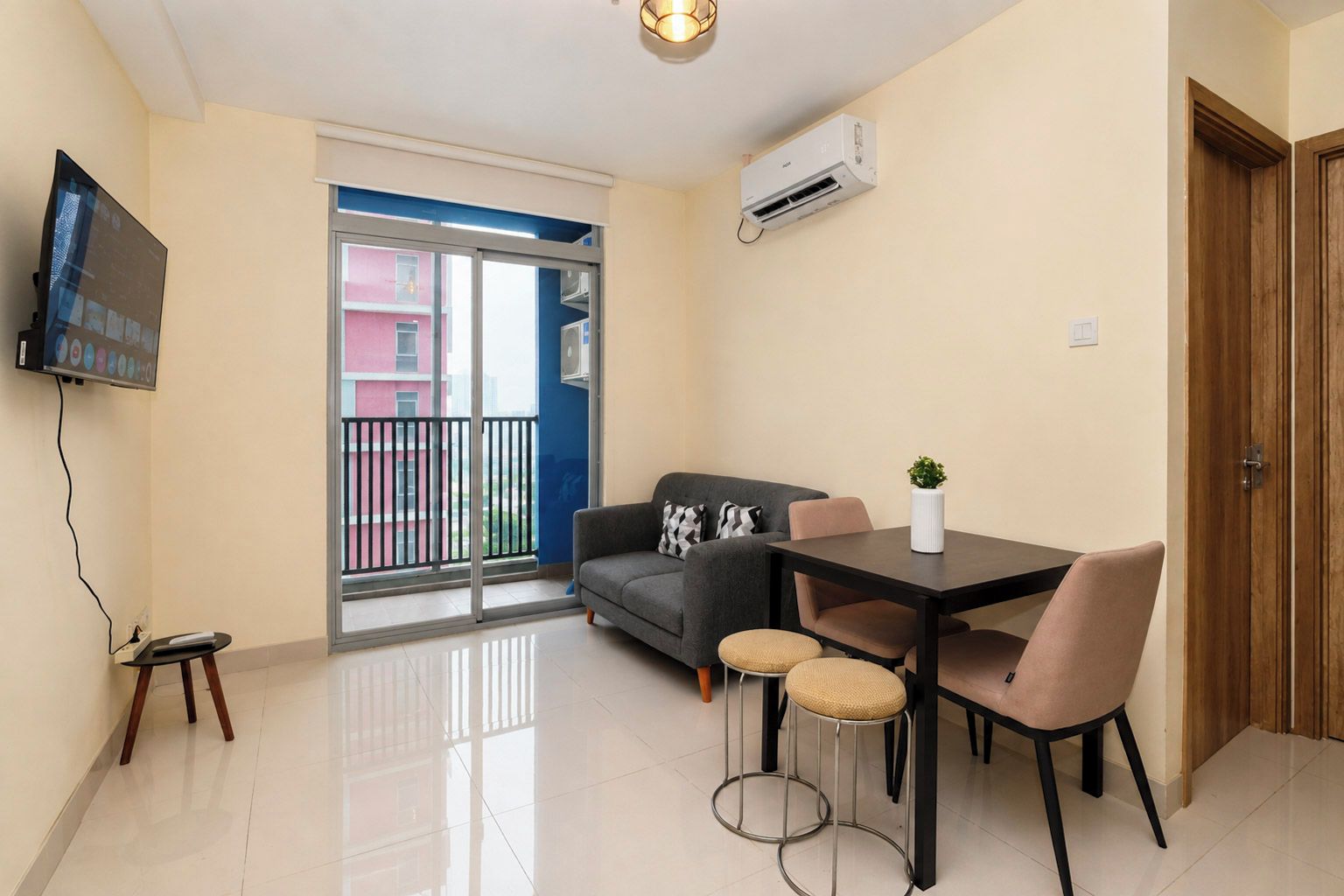 A modern living room with a sofa, a dining table with chairs, a TV mounted on the wall, and an air conditioning unit.