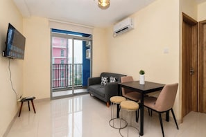 A modern living room with a sofa, a dining table with chairs, a TV mounted on the wall, and an air conditioning unit. - Warm And Sleek 1Br At Apartment Pejaten Park Residence (Jakarta)