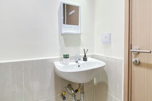 A bathroom with a white sink, a small potted plant, a mirror, and a door with a handle. - Warm And Peaceful 2Br At Apartment Meikarta District 1 (Cikarang)