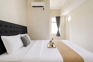 Habitación de hotel con una cama, cabecero, equipo de aire acondicionado, ventana con cortinas y bandeja con planta y velas.