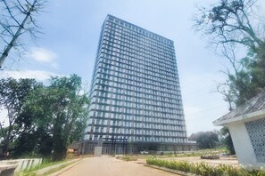 A tall, modern building with a glass facade, surrounded by greenery and a paved area in front. - Good And Practical Studio Room At Apartment Sudimara Forestwalk (Tangerang)