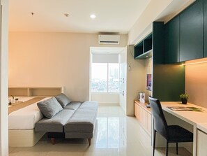 A compact living space with a sofa, a desk, and a chair. There is a window with a view of buildings outside. - Good And Warm Studio Room At Apartment Grand Kamala Lagoon (Bekasi)