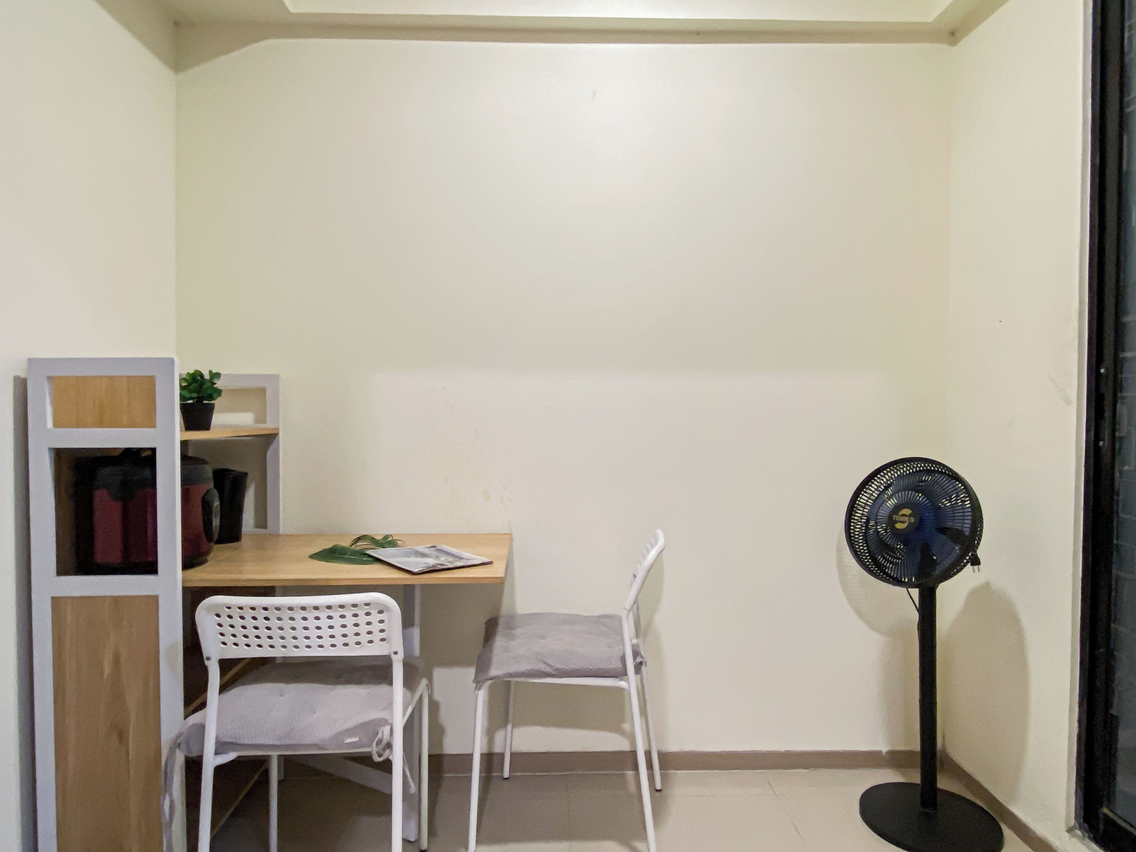 Une petite pièce comprenant un bureau, deux chaises, un ventilateur sur pied et une étagère avec une plante.