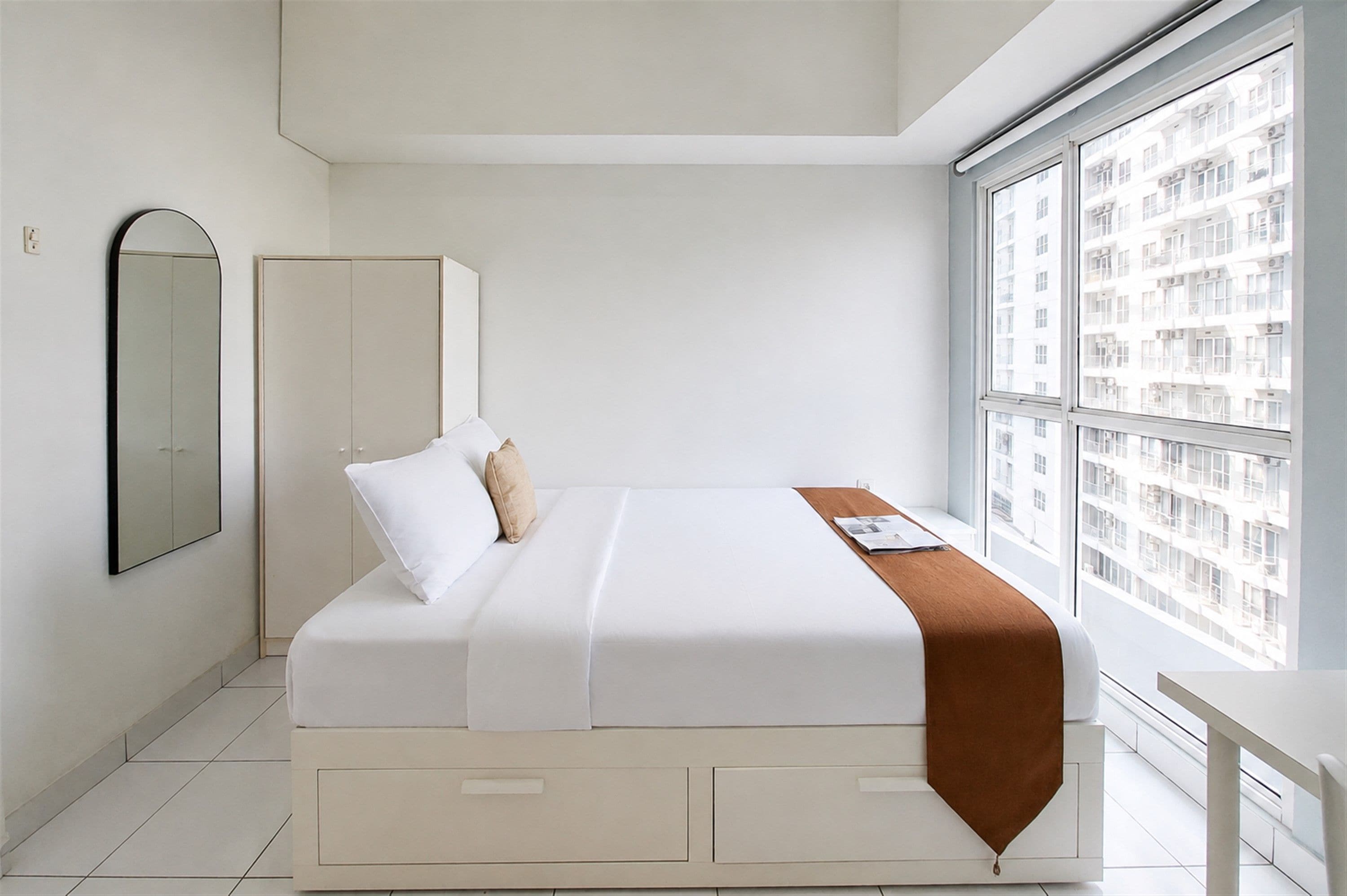 Quarto moderno com cama, guarda-roupa, janela e vista para prédios, além de mesa de cabeceira.