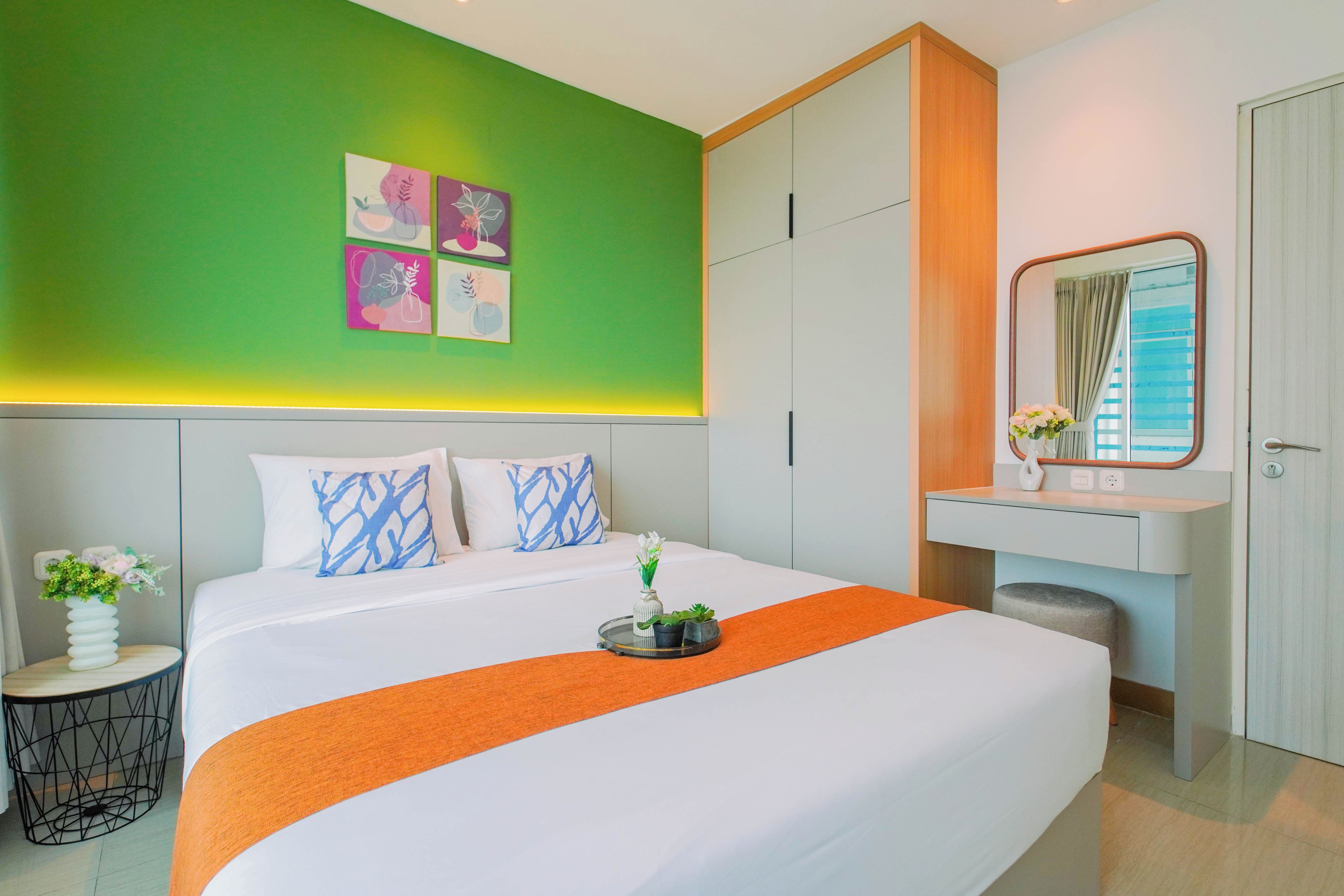 Habitación de hotel con cama, mesita de noche con un florero, espejo y pared verde con obras de arte.