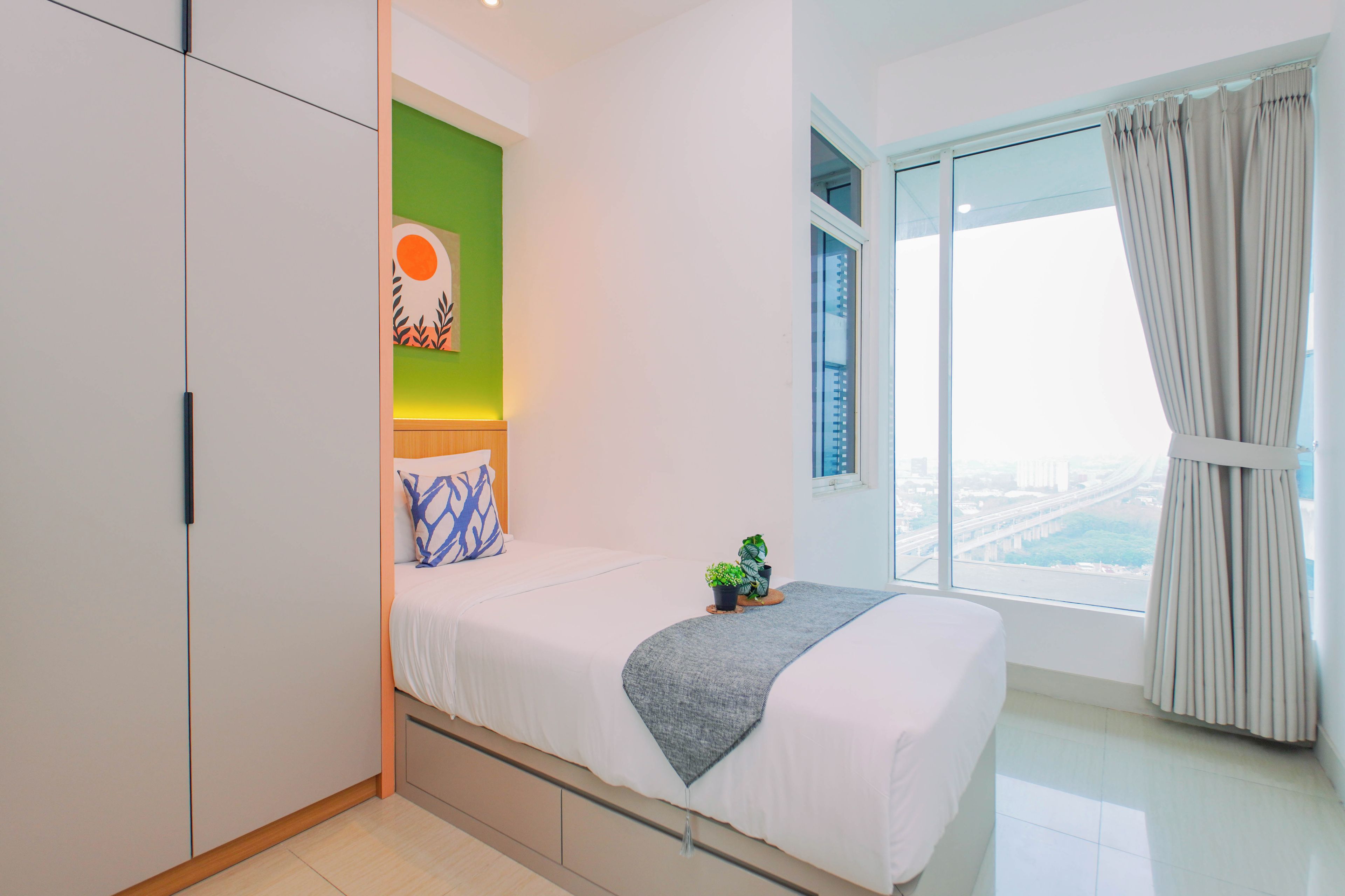 Un dormitorio con cama, pared de acento verde, ventana con cortinas y vistas a un paisaje urbano.