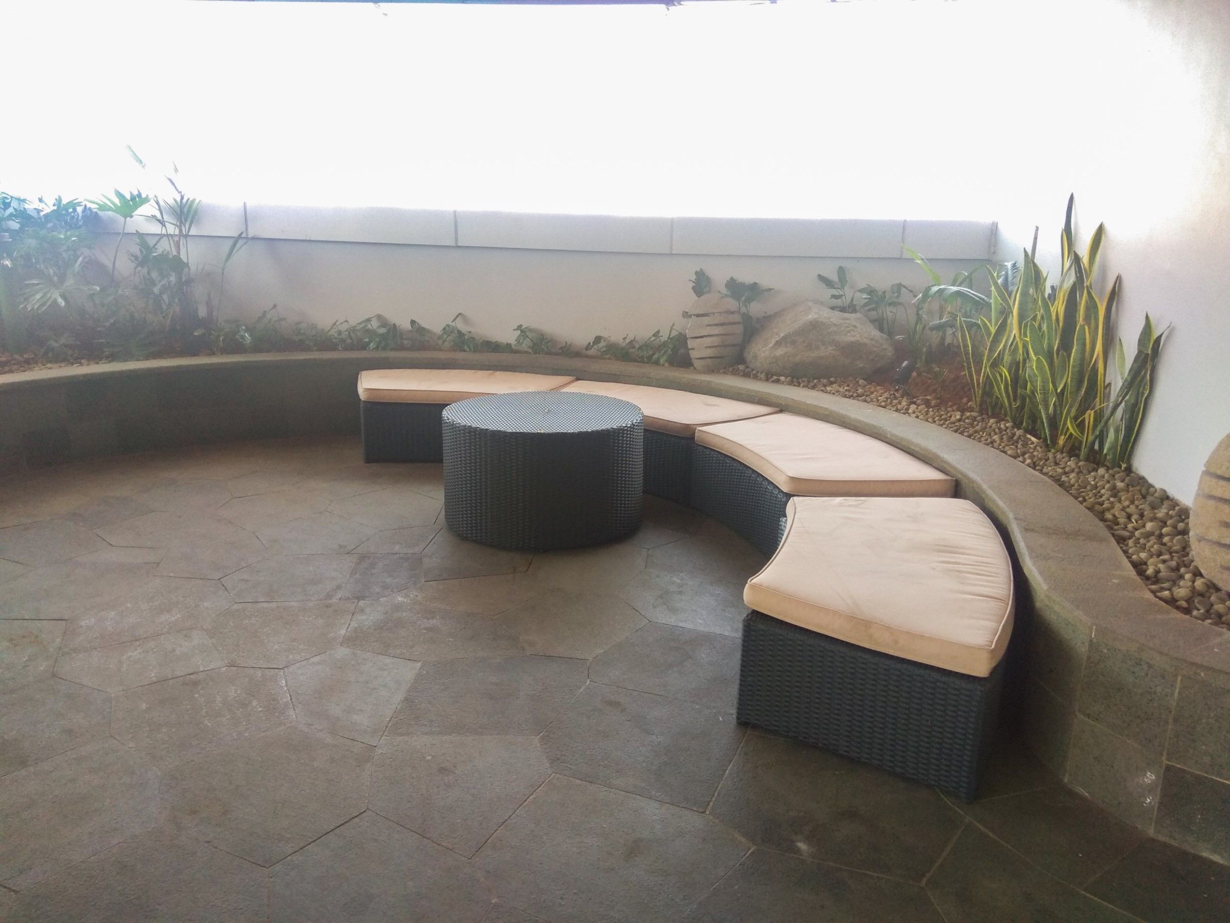 Un área de estar al aire libre con curvas, con bancos acolchados y un otoman redondo, rodeada por un bajo muro de piedra y diversas plantas.