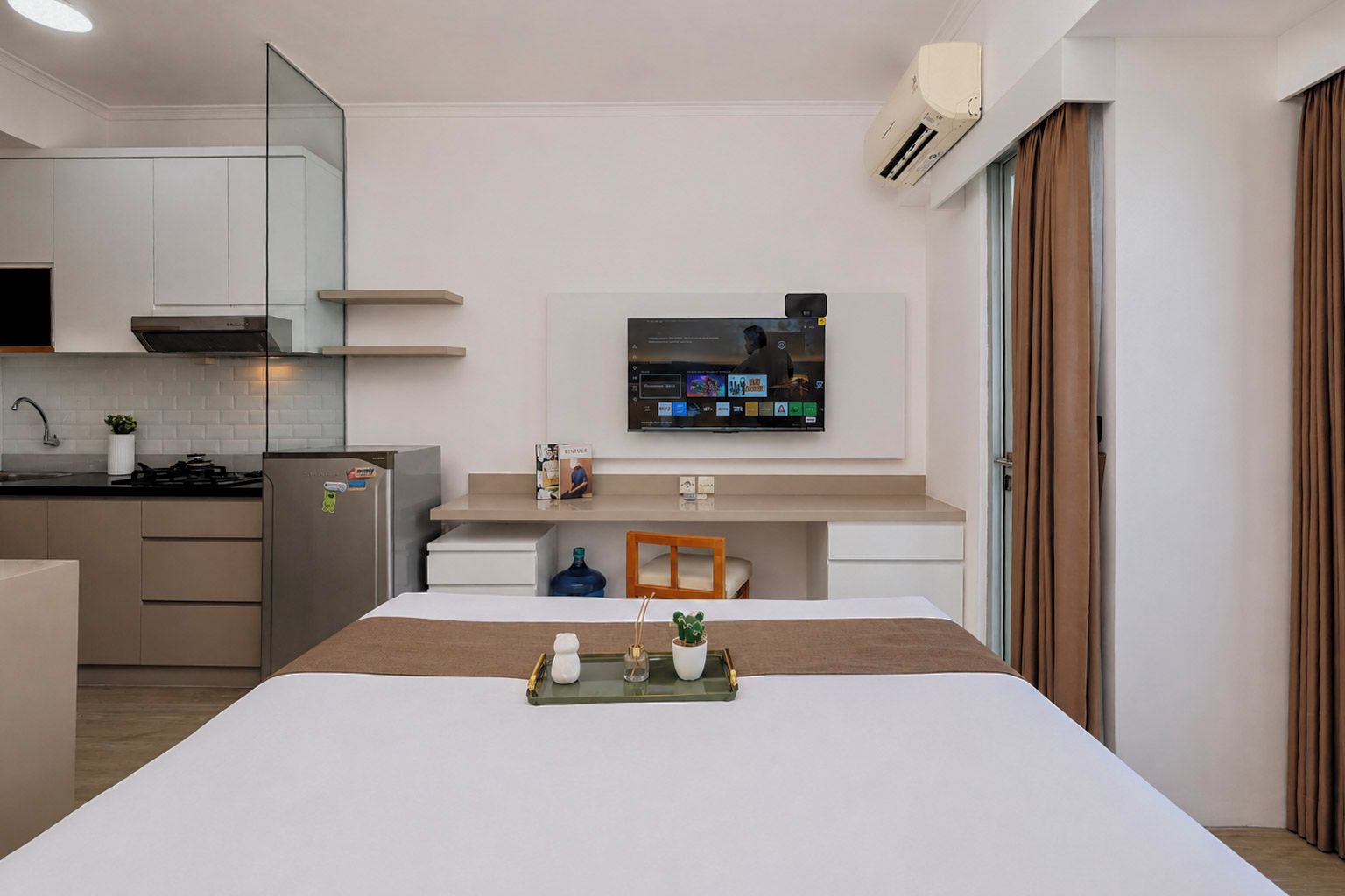 A modern hotel room with a kitchen area, a flat-screen TV mounted on the wall, and a bed with a tray on it.
