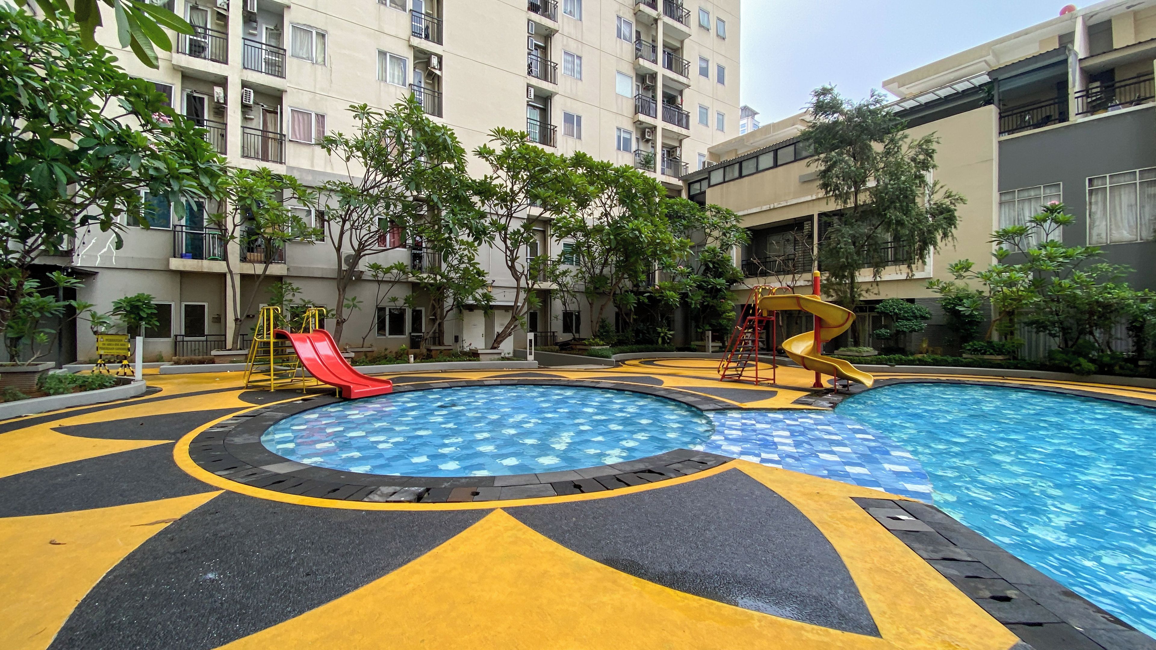 A swimming pool with a slide and a spiral slide, surrounded by a paved area with yellow and black markings, and residential buildings in the background.