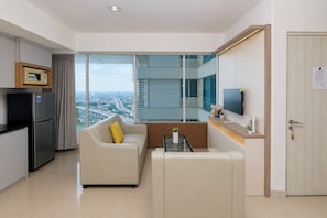 A modern hotel room with a kitchenette, a sofa, a coffee table, a TV, and a large window offering a city view. - Comfortable 2Br At Apartment Grand Kamala Lagoon (Bekasi)