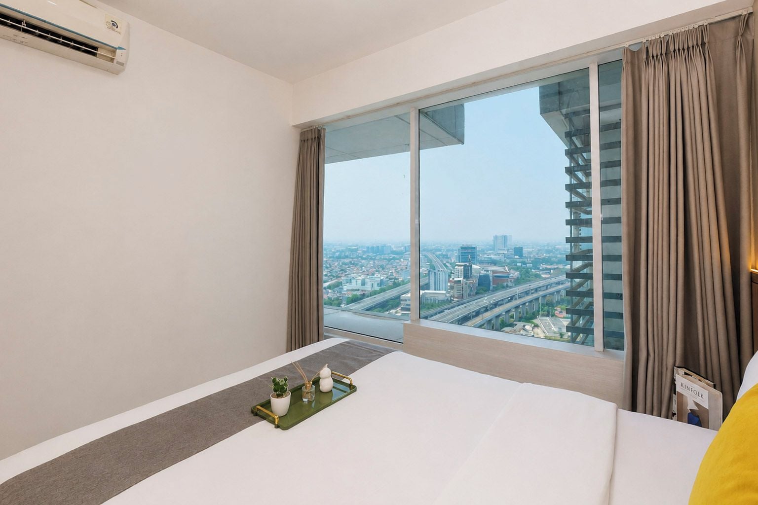 Ein Hotelzimmer mit großem Fenster, einem Bett, einem Nachttisch mit Tablett und einer Klimaanlage.
