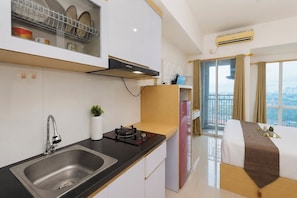 A modern kitchen with a sink, stove, and dish rack. There is a small plant on the counter and a window with curtains. - Good Homey Studio Room At Apartment Grand Dhika City (Bekasi)