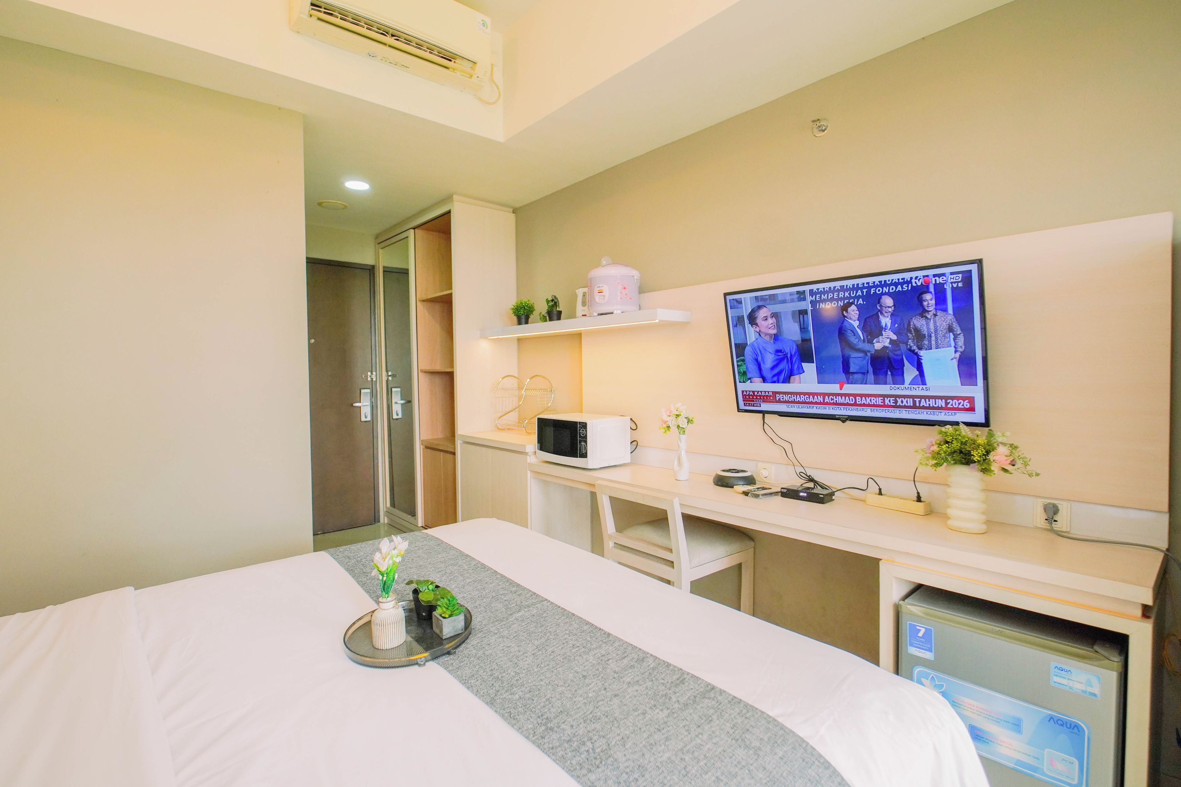A hotel room with a bed, a TV mounted on the wall, a microwave, a small shelf with plants, and a water dispenser.