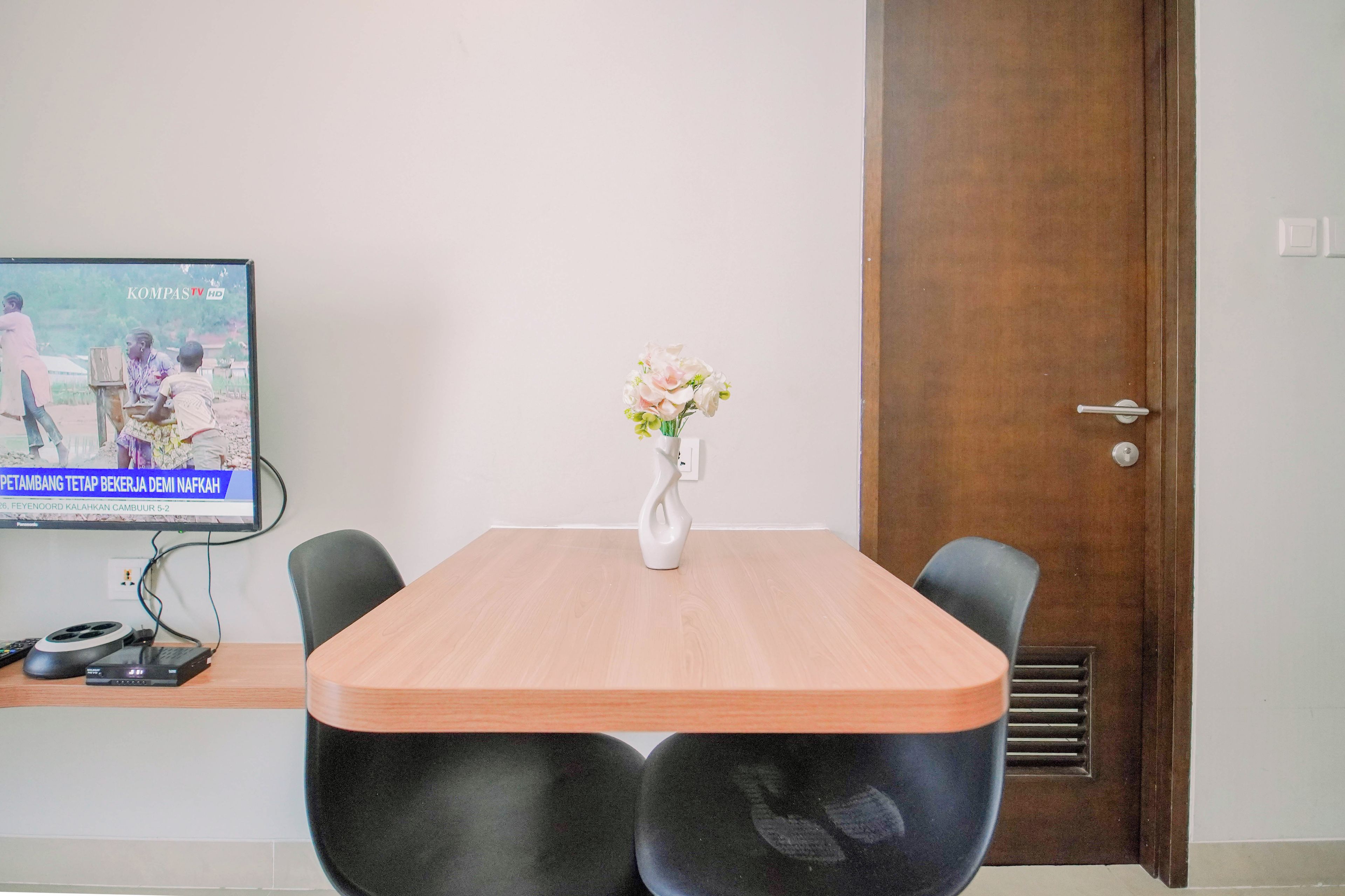 A conference room with a wooden table, black chairs, a white vase with flowers, and a television displaying a news program.