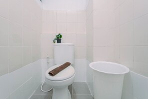 A small, white bathroom with a toilet, a white trash bin, and a potted plant on the back of the toilet. - Good And Homey 2Br At Apartment Kemang View (Bekasi)