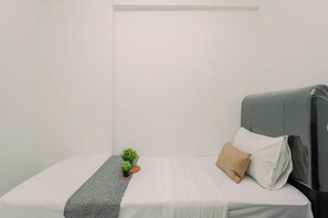 Uma cama de solteiro com cabeceira cinza, roupa de cama branca, uma pequena planta em vaso e um cobertor cinza.