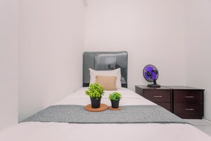 A neatly made bed with a grey blanket, two potted plants, a single pillow, and a bedside table with a fan. - Good And Homey 2Br At Apartment Kemang View (Bekasi)