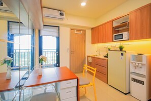A compact kitchen with wooden cabinets, a microwave, a refrigerator, and a small dining table with a chair. - Pleasant Warm Studio Room At Apartment Meikarta District 2 (Cikarang)