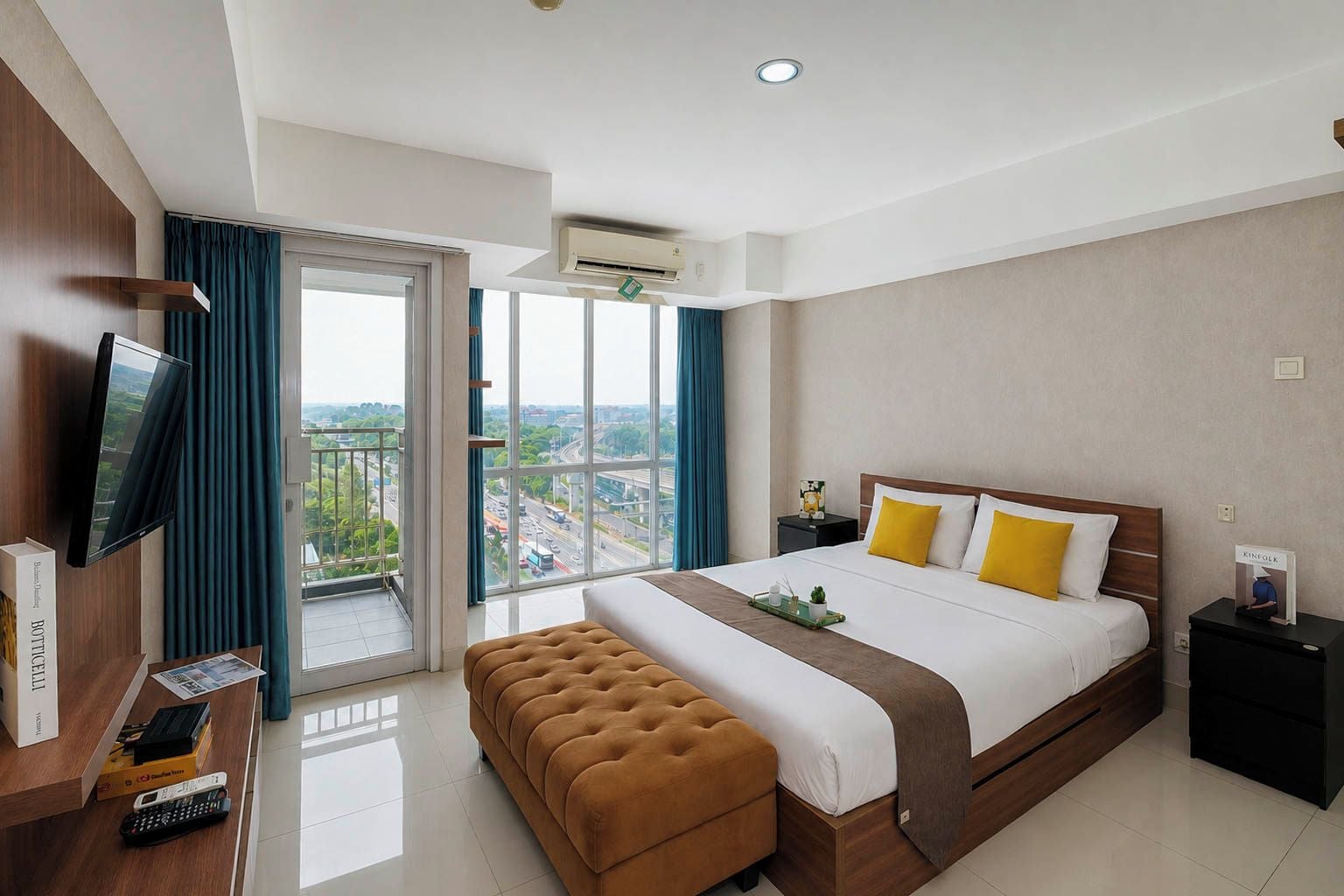 A hotel room with a bed, a television, a desk with a remote control, a bench, and a view of the city through large windows.