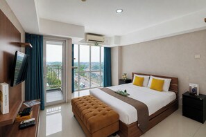 A hotel room with a bed, a television, a desk with a remote control, a bench, and a view of the city through large windows. - Spacious And Warm Studio At Apartment H Residence (Jakarta)