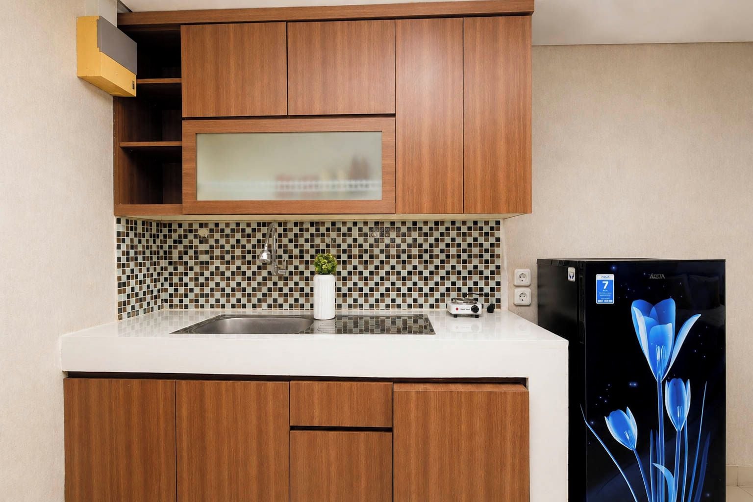 A kitchen with wooden cabinets, a white countertop, a tiled backsplash, a stainless steel sink, a small potted plant, and a black refrigerator.