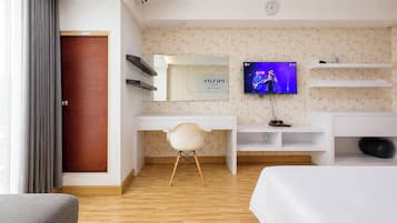 A hotel room with a wooden floor, a white desk, a chair, a bed, a television, and a wardrobe.