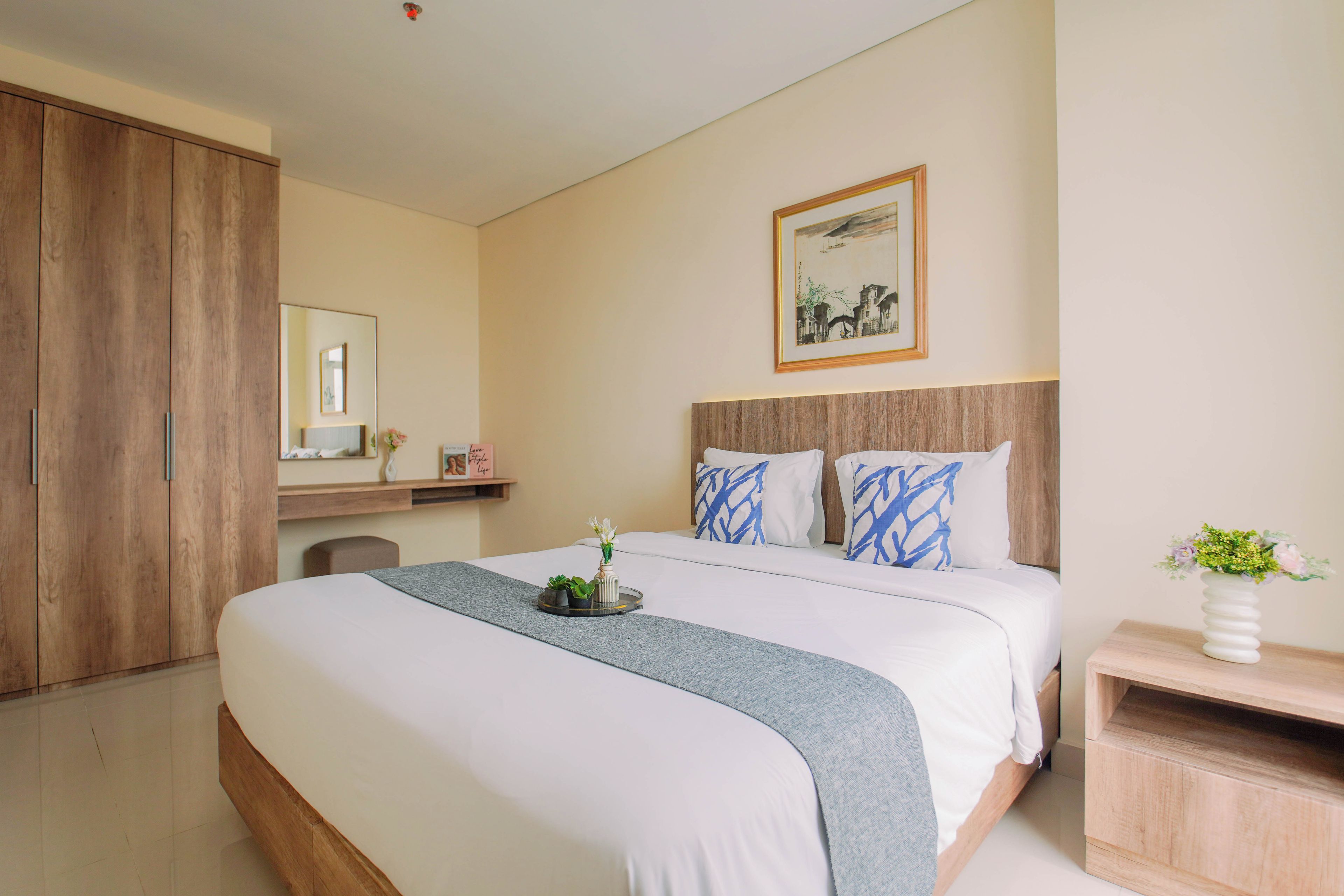 Una habitació d'hotel amb un llit gran, un armari de fusta, una taula de nit amb un vas de flors i un quadre a la paret.