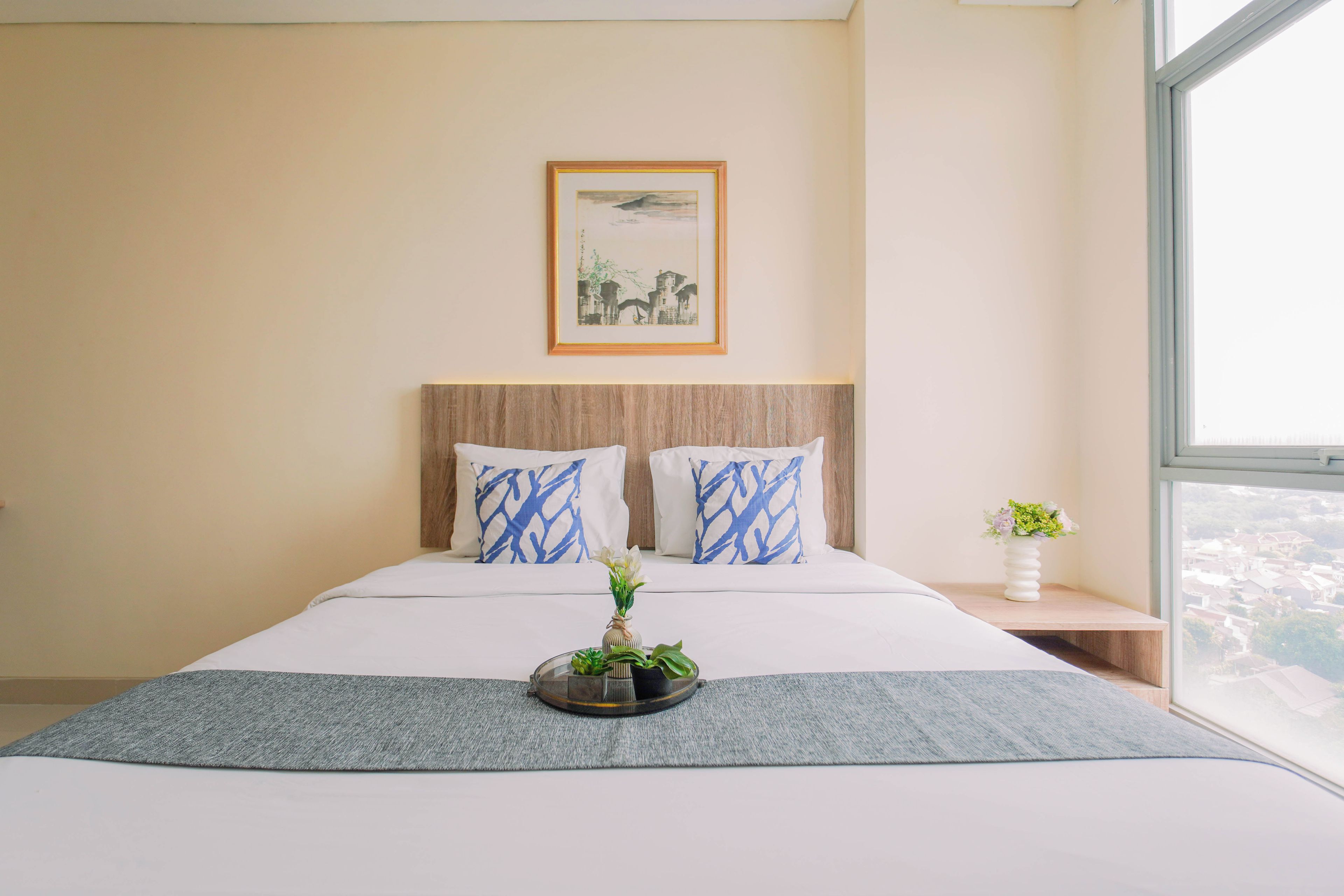 A hotel room with a large bed, a headboard, a framed picture on the wall, a bedside table with a vase, and a view through the window.