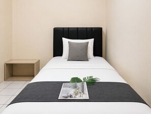 Uma cama de solteiro com cabeceira preta, roupa de cama branca e travesseiro cinzento. Uma revista está colocada sobre a cama.