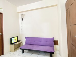 Quarto com um sofá roxo, uma televisão sobre um suporte e uma parede branca.