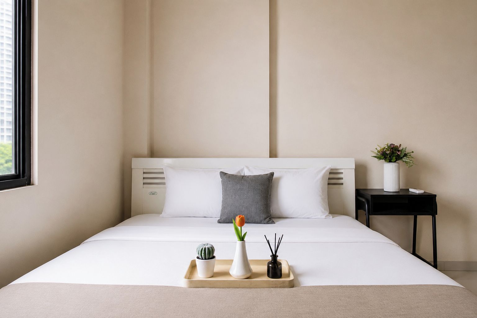 Una cama bien hecha con ropa de cama blanca, un almohadón gris, un almohadón blanco, una pequeña planta en maceta, una bandeja con una planta en maceta y una vela, y una mesita con una planta y un control remoto.