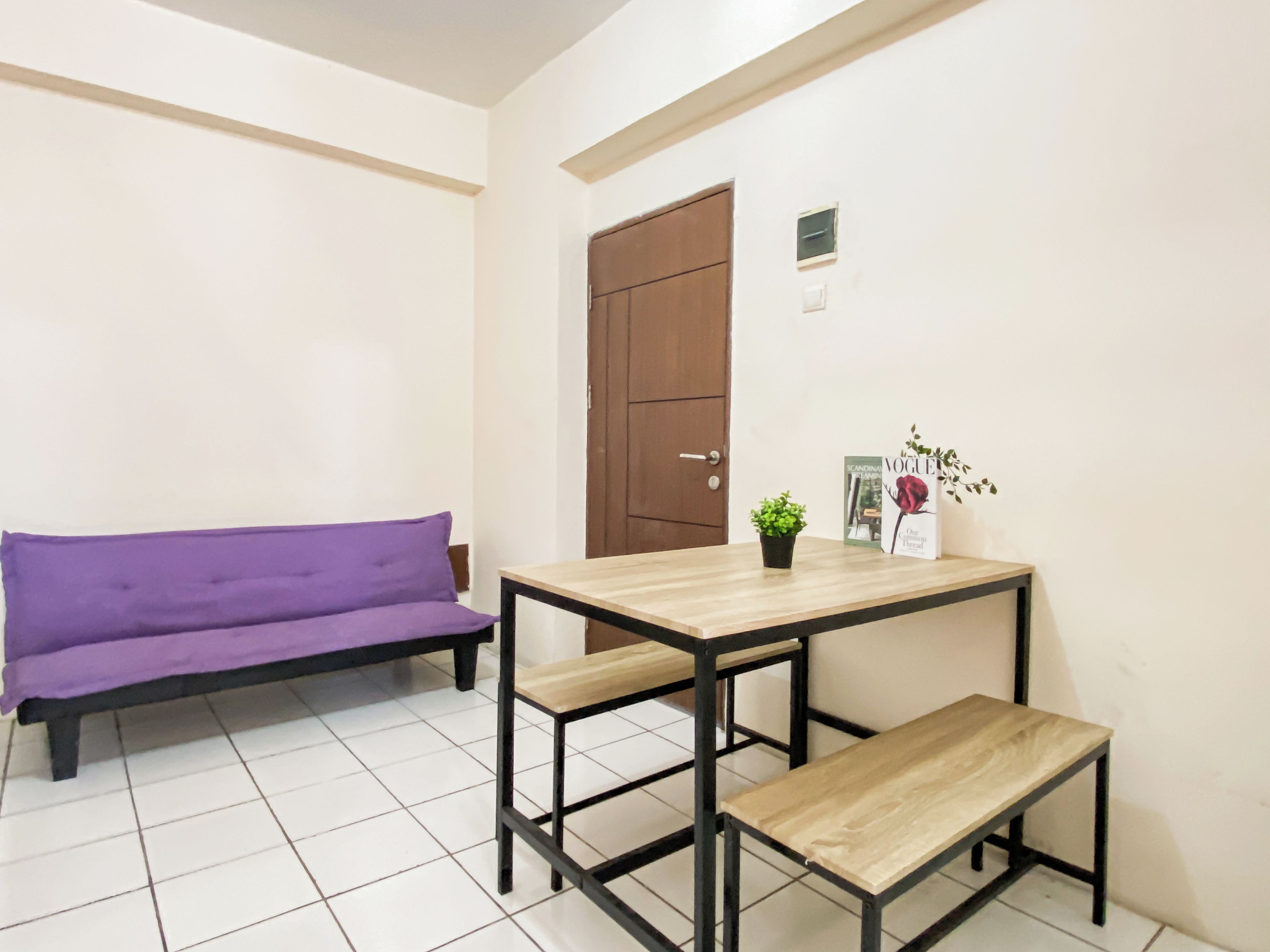 Una habitación con un sofá morado, una mesa de madera con bancos, una planta pequeña en maceta y una puerta.