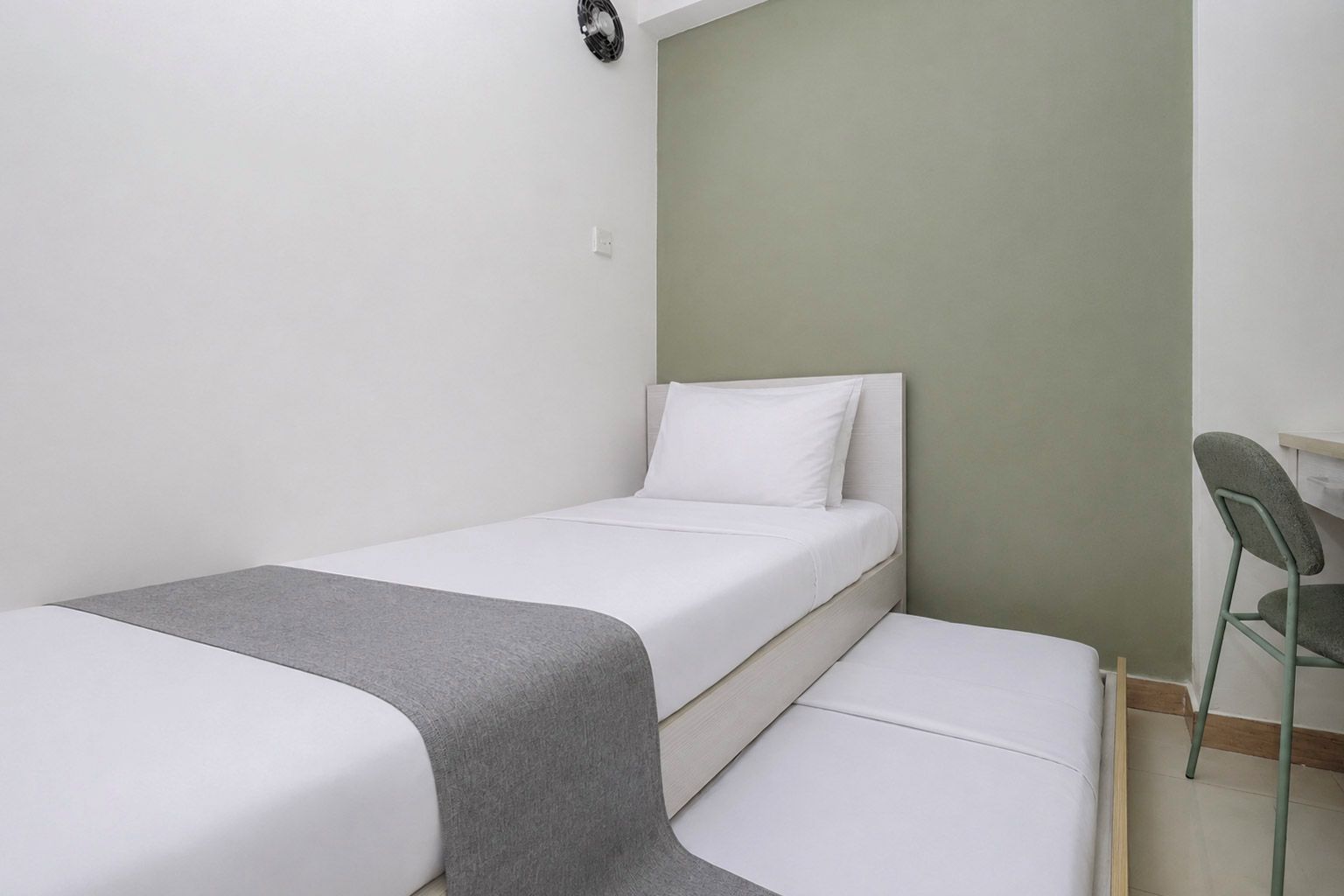 Habitación individual con una cama, un escritorio, una silla, una mesita de noche pequeña y un ventilador de pared.