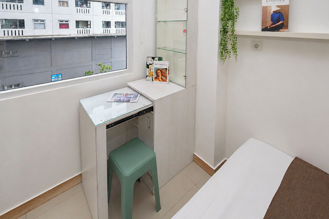 A small, clean room with a window, a desk, a green stool, a bed, and a shelf with a book.