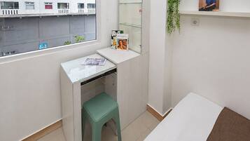 A small, clean room with a window, a desk, a green stool, a bed, and a shelf with a book.