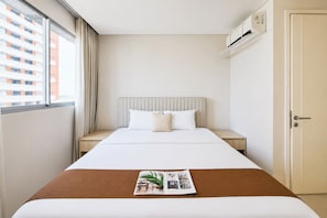 A hotel room with a bed, a nightstand, a window with curtains, an air conditioning unit, and a door.