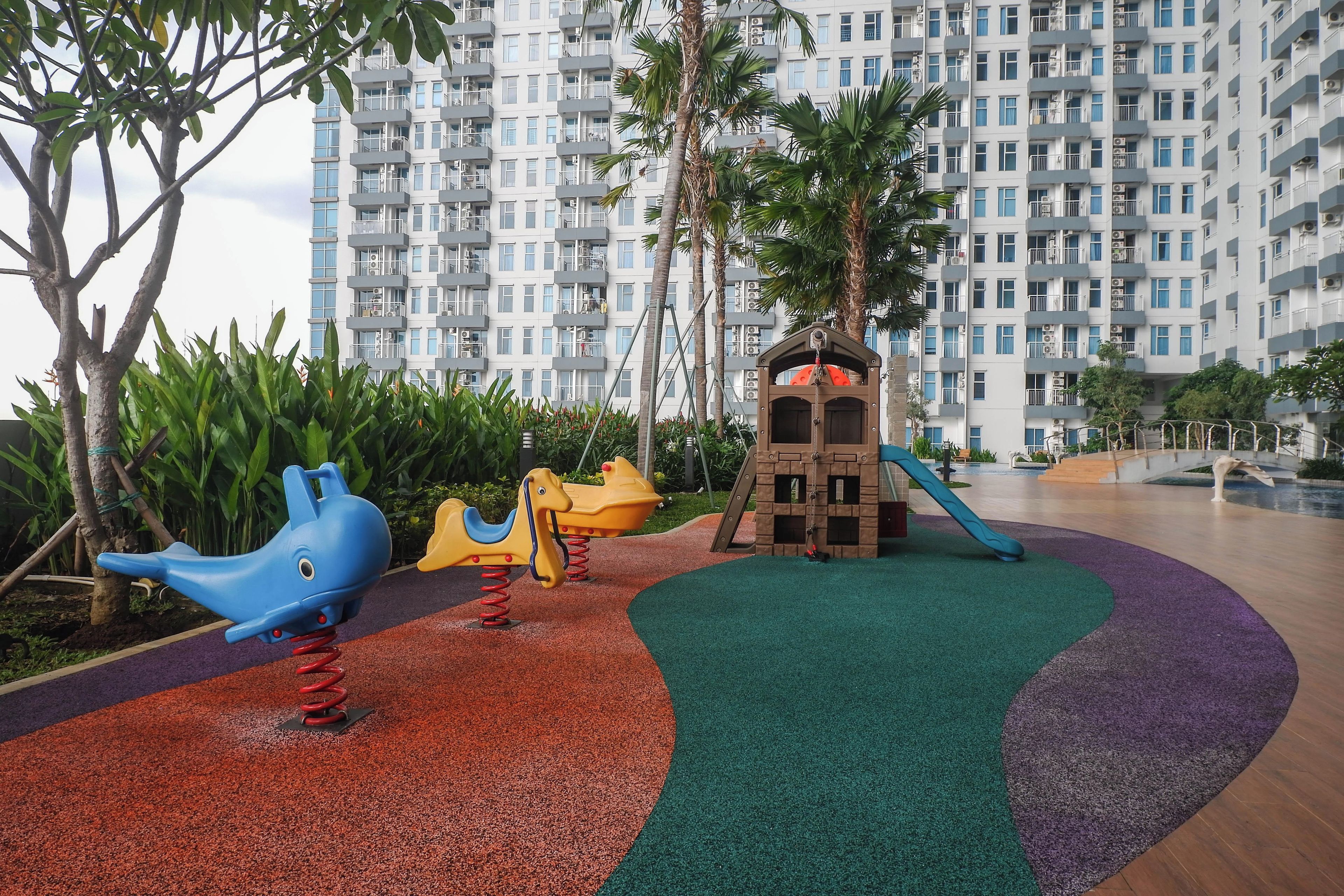 A playground with a slide and climbing structures, surrounded by a landscaped area with trees and a building in the background.