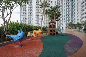 Ein Spielplatz mit Rutsche und Klettergerüsten, umgeben von einer Grünfläche mit Bäumen und einem Gebäude im Hintergrund.
