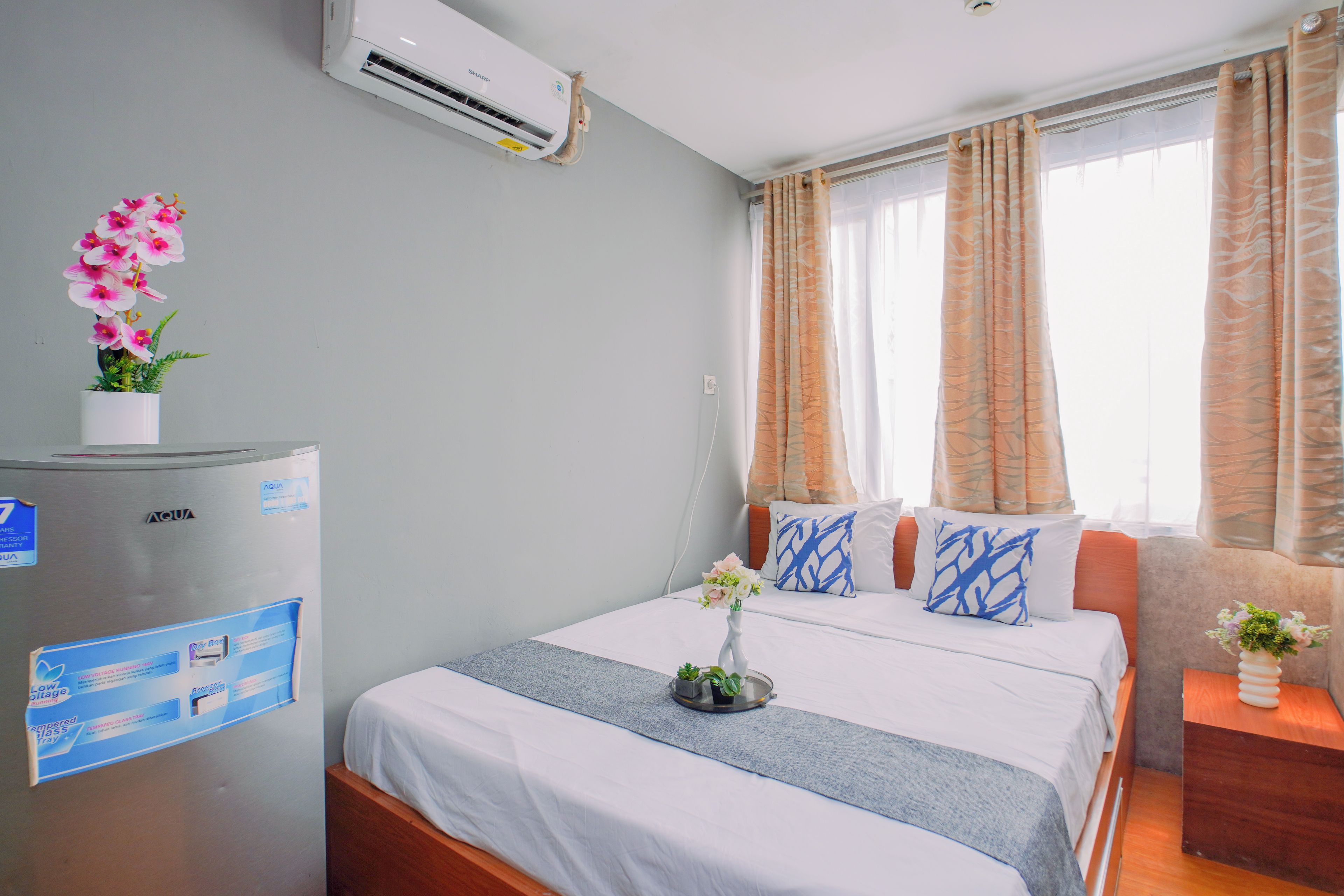 Quarto de hotel com cama, mesa de cabeceira, geladeira e janela com cortinas.