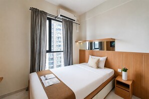 A hotel room with a bed, a bedside table, a nightstand, a window with curtains, and an air conditioning unit. - Modern Look Studio At Apartment Sky House Alam Sutera (Tangerang)