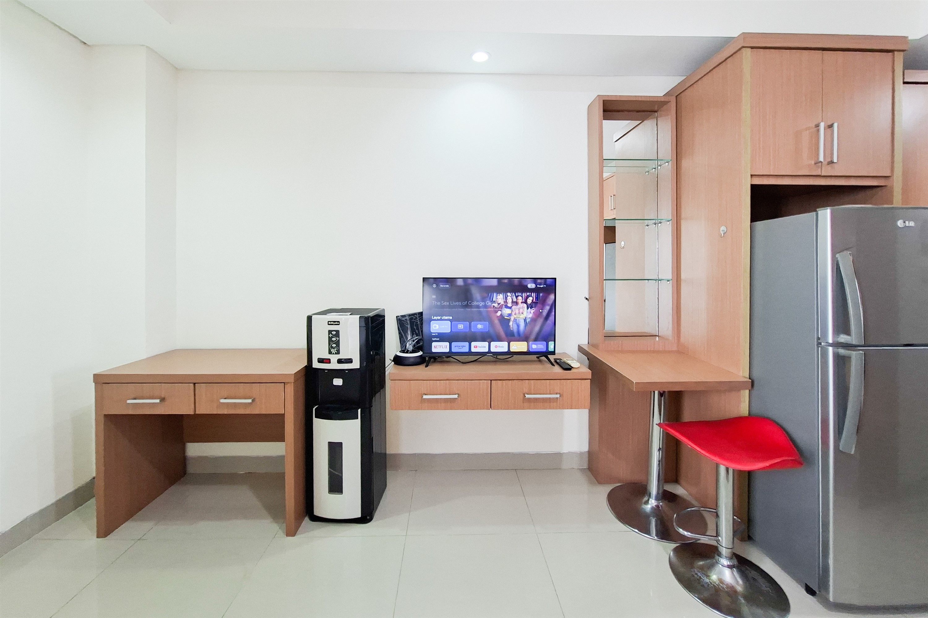Una kitchenette compacta con refrigerador, cafetera, computadora de escritorio y televisor mostrando un programa.