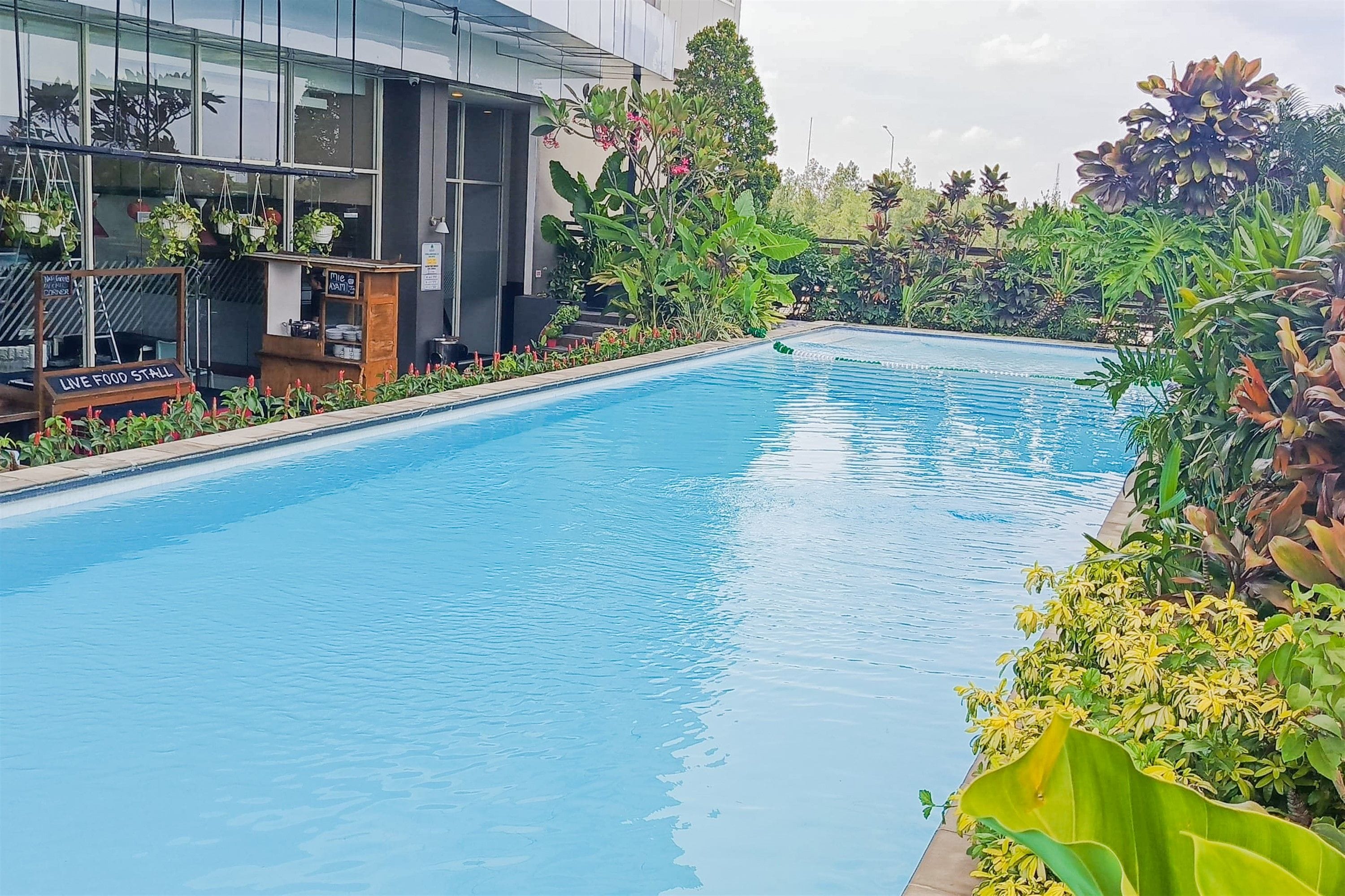 A swimming pool with a sign for a live food stall, surrounded by lush greenery and a modern building in the background.