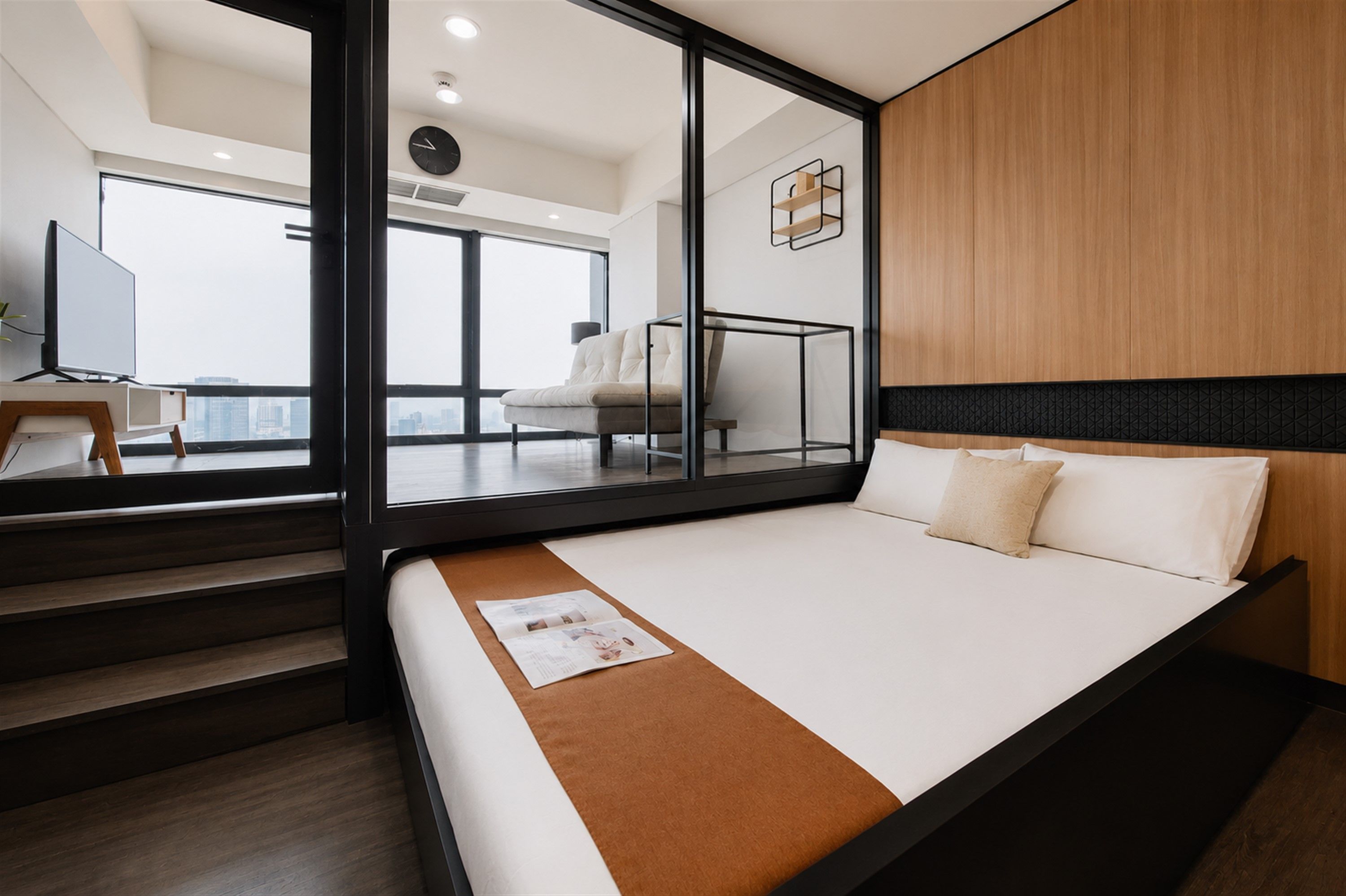 Una habitación de hotel moderna con una cama grande, cabecera de madera, separador de vidrio y vistas a la ciudad.