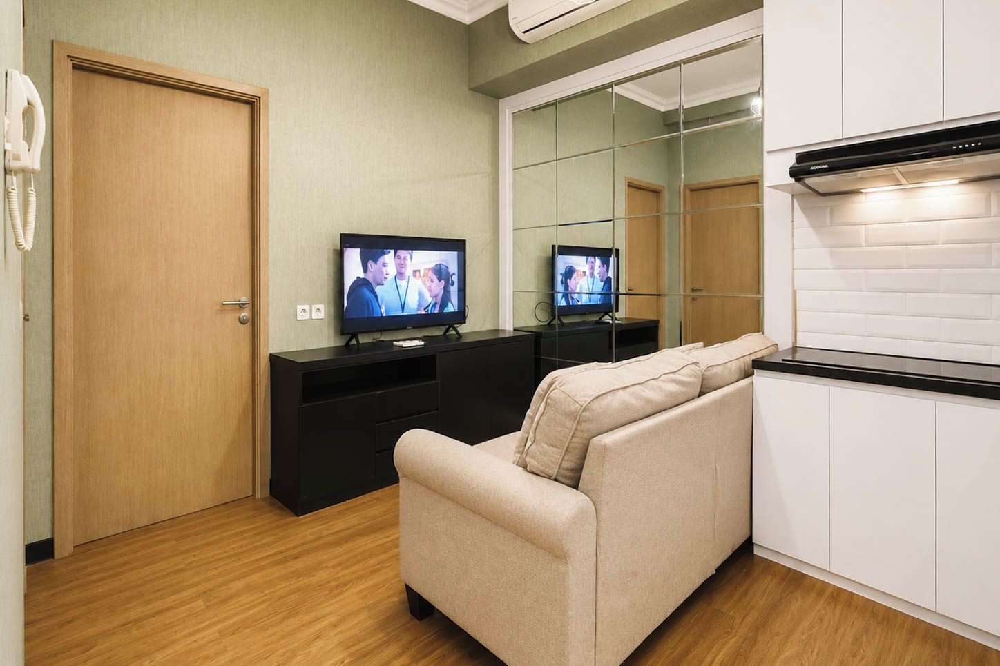 A compact living space with a sofa, a television, a wooden door, and a kitchen area with white cabinets and a black countertop.