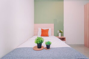 Ein ordentlich bezogenes Bett mit einem weiß-orangenen Kopfkissen, zwei kleine Topfpflanzen und ein Nachttisch aus Holz mit einem Blumenstrauß.