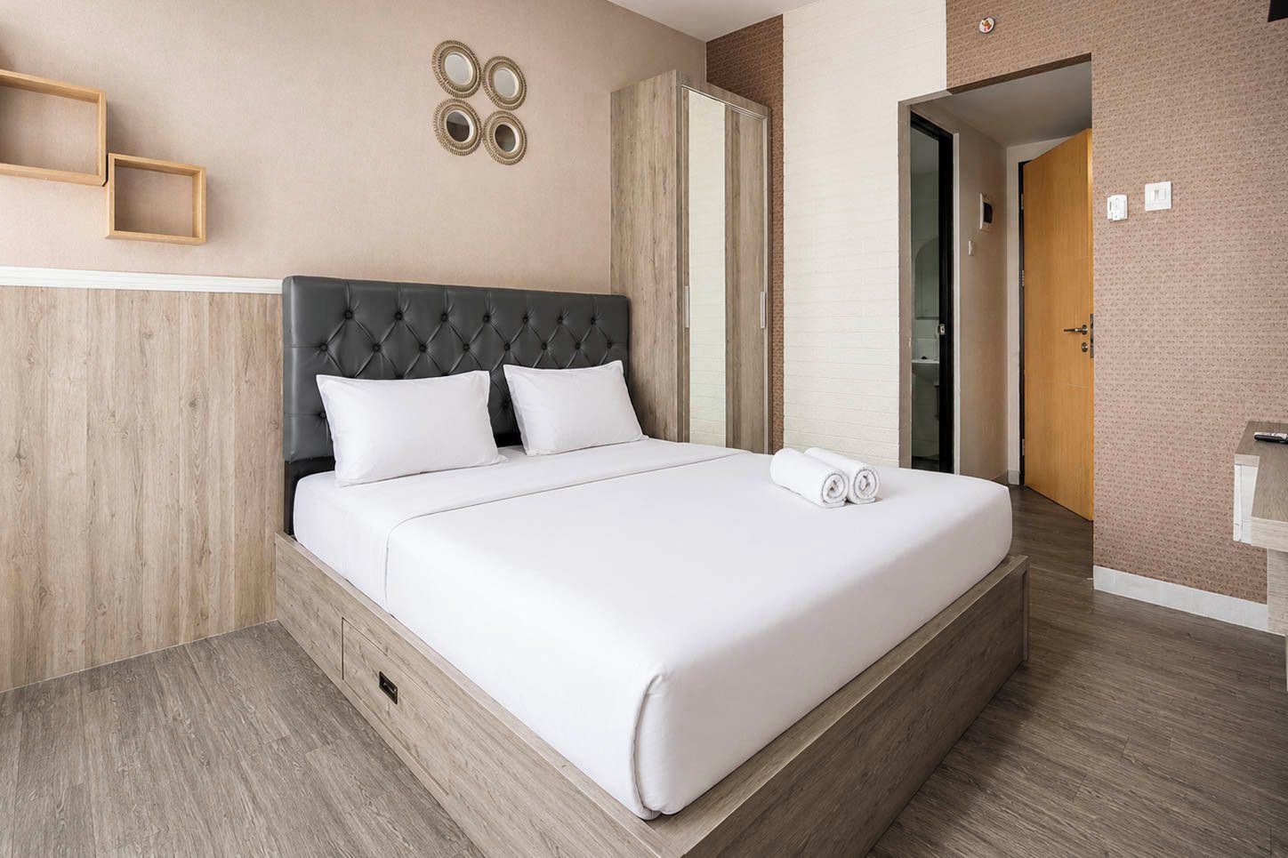 A neatly made bed with a tufted headboard, a wooden floor, and a wall with a mirror and wall art.