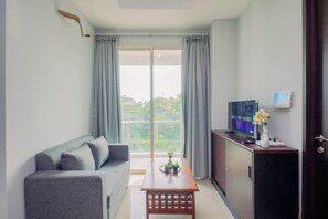 A modern living room with a grey sofa, a wooden coffee table, a TV on a cabinet, and a view of greenery through large windows. - Cozy And Fully Furnished 1Br At Apartment Citralake Suites (Jakarta)